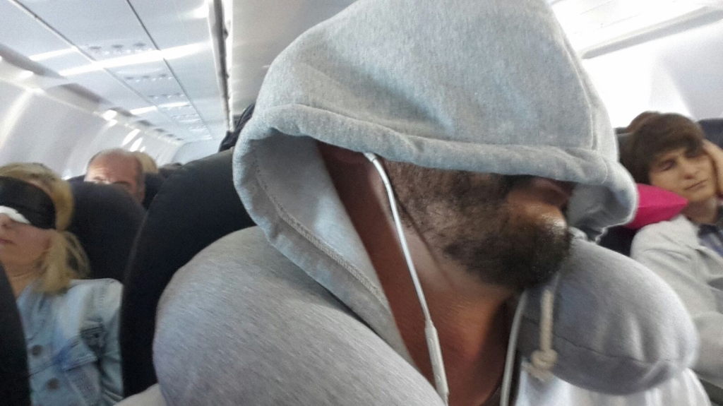 Mann mit Kapuzenpullover und Nackenkissen schläft im Flugzeug auf dem Rückflug von Samos