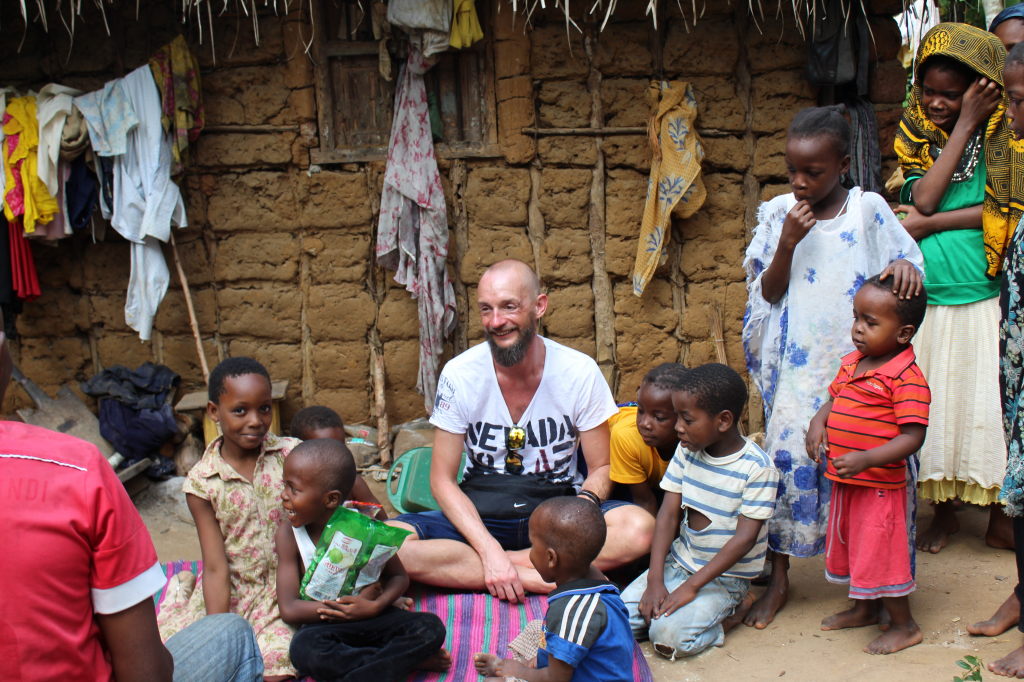 Reisender sitzt lachend mit Dorfkindern vor Lehmhaus in Mchangani, Sansibar