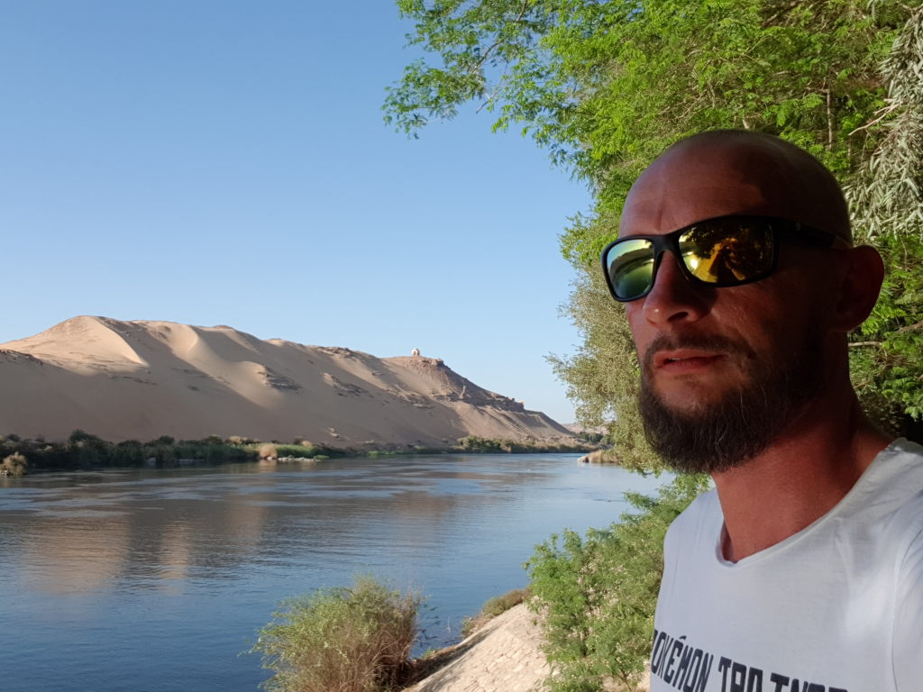 Mann mit Sonnenbrille macht Selfie am Nil in Assuan mit Sanddünen im Hintergrund