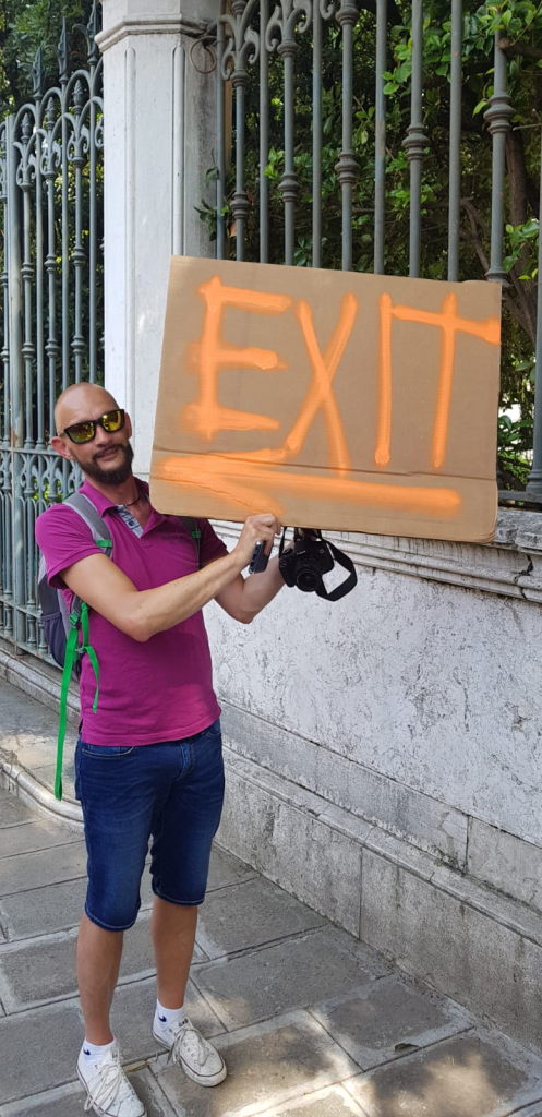 Mann mit Sonnenbrille hält EXIT-Schild aus Pappe vor Metallzaun in Venedig