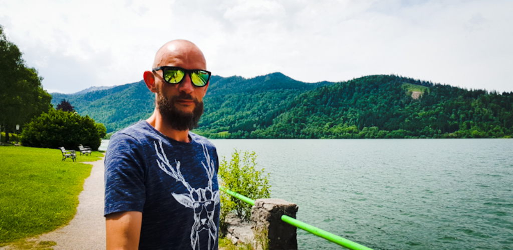 Mann mit Sonnenbrille am Schliersee-Ufer mit bewaldeten Bergen im Hintergrund