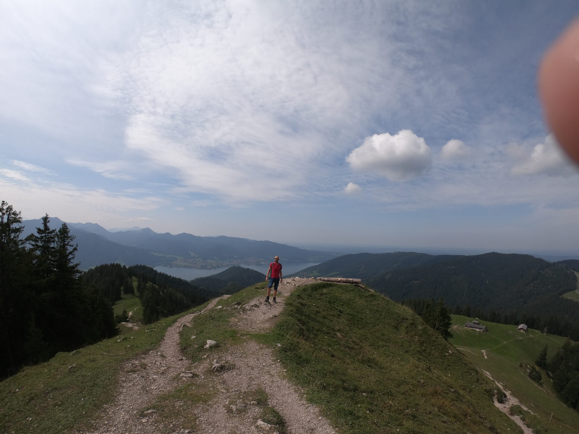 Wanderer auf dem schmalen Gratrücken des Baumgartenschneid mit Tegernsee-Panorama im Hintergrund und Almhütte unten rechts