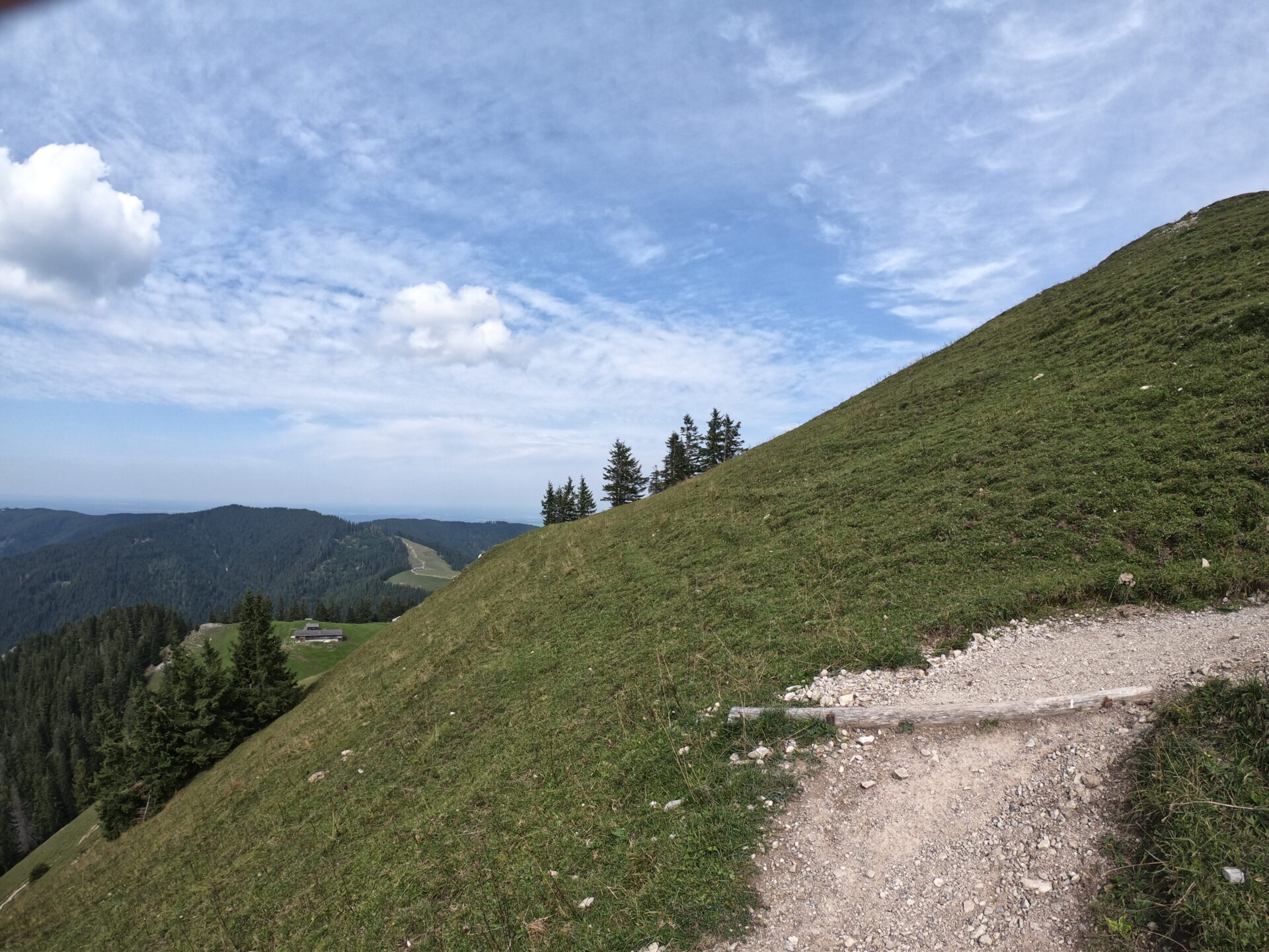 Schmaler Wanderweg am steilen Grashang des Baumgartenschneid mit Blick auf einen Almhof und bewaldete Berge im Hintergrund