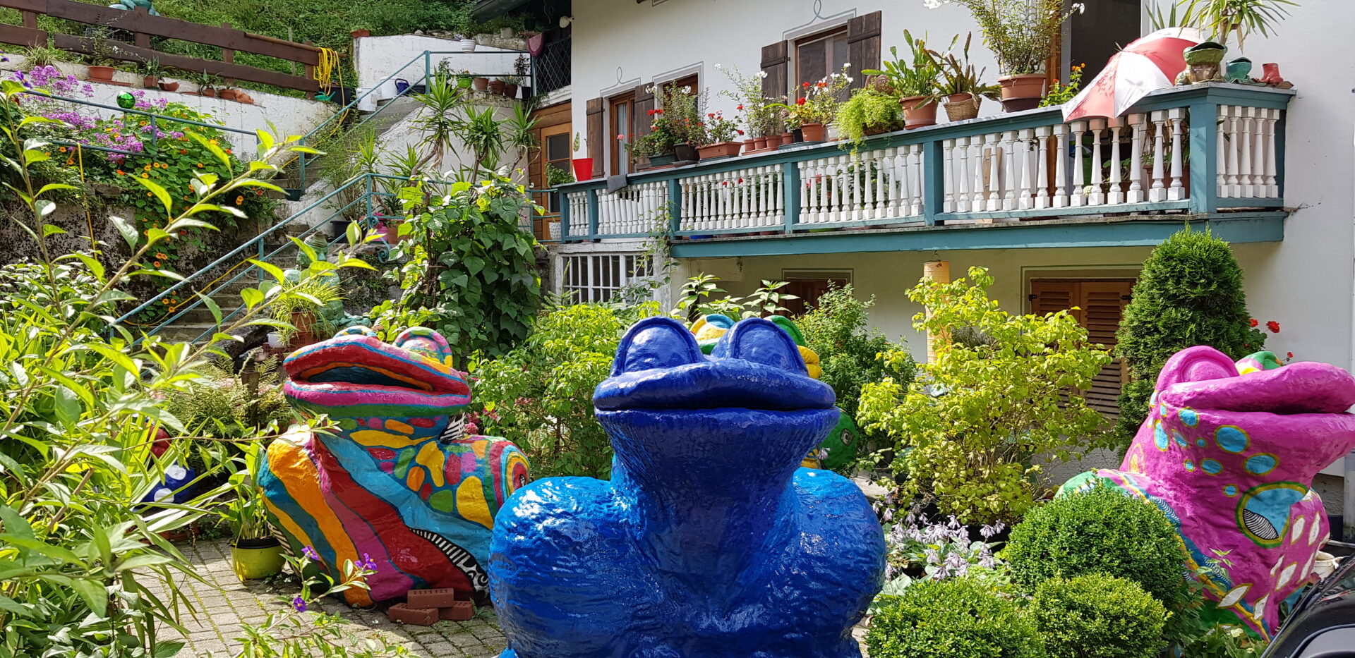 Bunte Froschskulpturen in einem üppig bepflanzten Garten in Tegernsee, im Vordergrund ein blauer Keramikfrosch