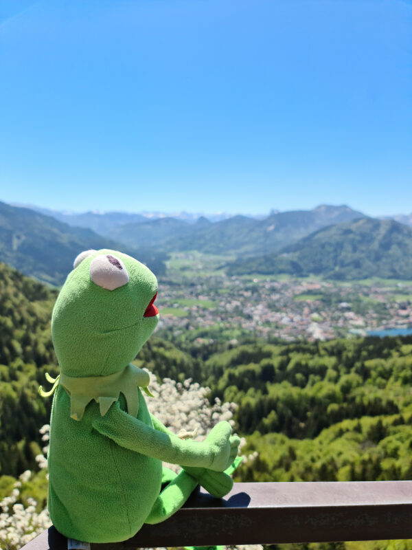 Kermit sitzt am Geländer der Riederstein Kapelle mit Blick auf den Tegernsee