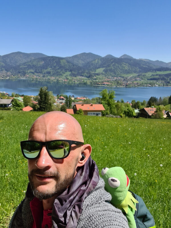 Sebastian mit Sonnenbrille und Kermit auf der Schulter, dahinter der Tegernsee und Berge