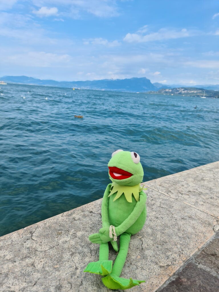 Kermit sitzt auf einer Steinmauer am Molo in Lazise mit dem blauen Gardasee und Bergen im Hintergrund