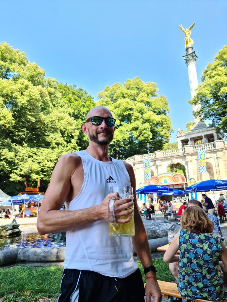 Sebastian mit Masskrug im Biergarten am Friedensengel München, goldene Engelstatue im Hintergrund, Sommer August 2020