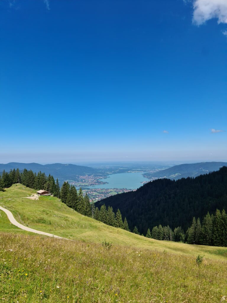 Ausblick vom Wallberg auf eine Almhütte, die Wiesen und den türkisen Tegernsee im Tal