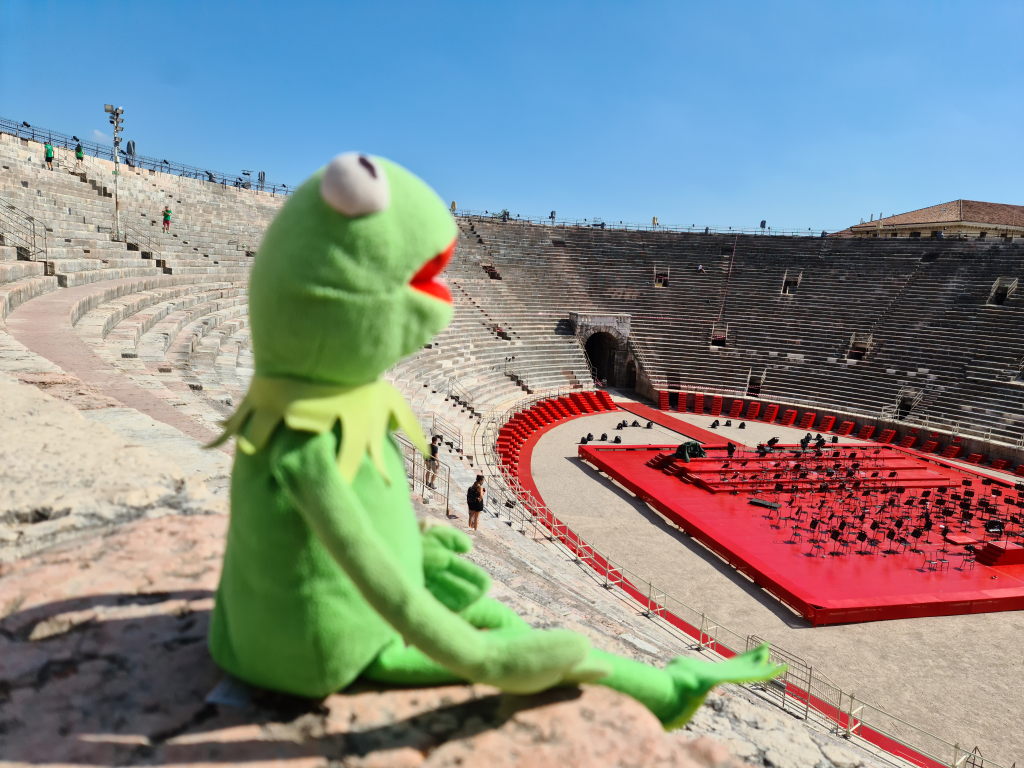 Kermit sitzt auf den Steinstufen der Arena di Verona und schaut auf die rote Orchesterbühne in der Mitte des Amphitheaters
