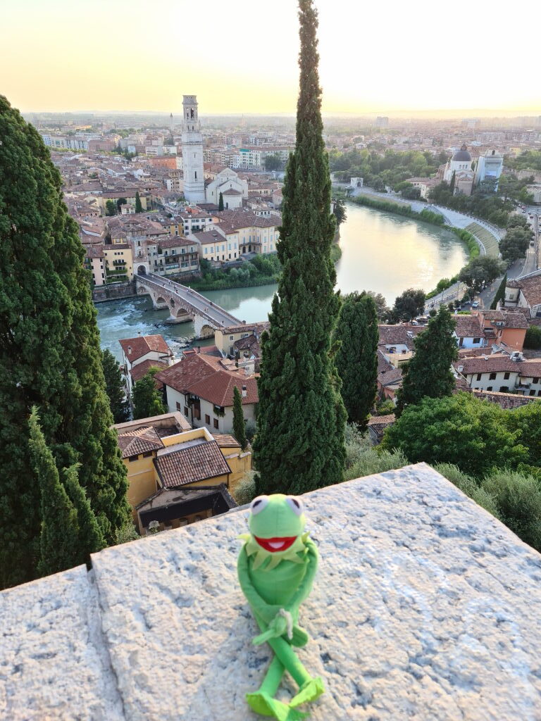 Kermit sitzt auf der Brüstungsmauer am Castel San Pietro mit Panoramablick auf Verona, Etsch und Zypressen beim Sonnenuntergang