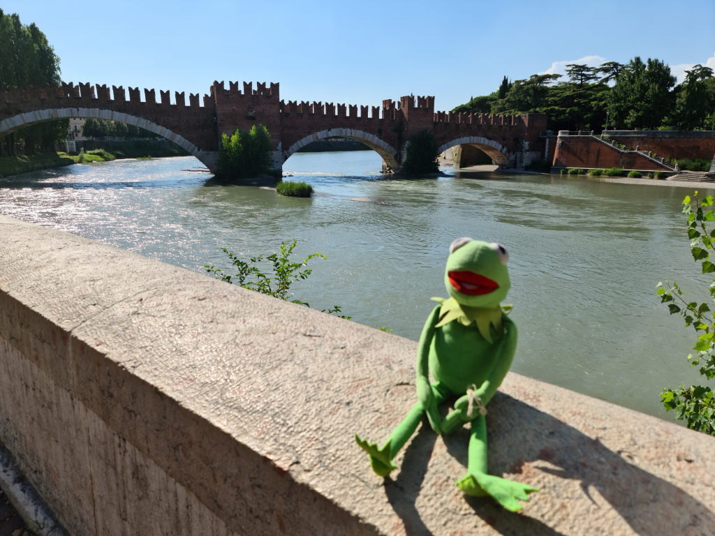 Kermit sitzt auf der Brüstungsmauer am Ponte Scaligero mit der Etsch und der mittelalterlichen Brücke im Hintergrund
