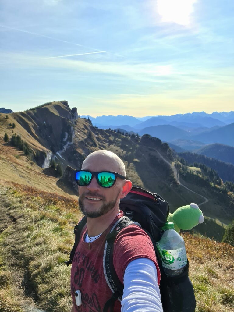 Selfie am Brauneck-Gipfel mit Kermit im Rucksack und Blick auf den Bergkamm im Oktober 2020