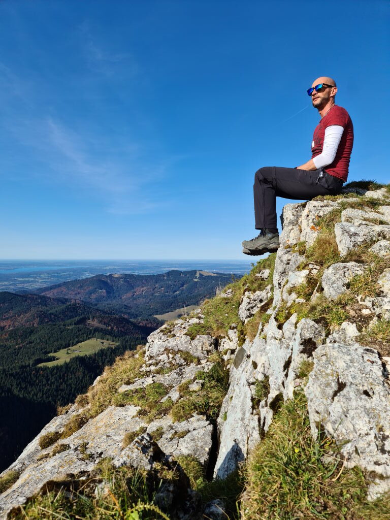 Wanderer sitzt auf Felsen am Kirchstein-Gipfel mit weitem Ausblick auf das Alpenvorland und den Starnberger See im Oktober 2020