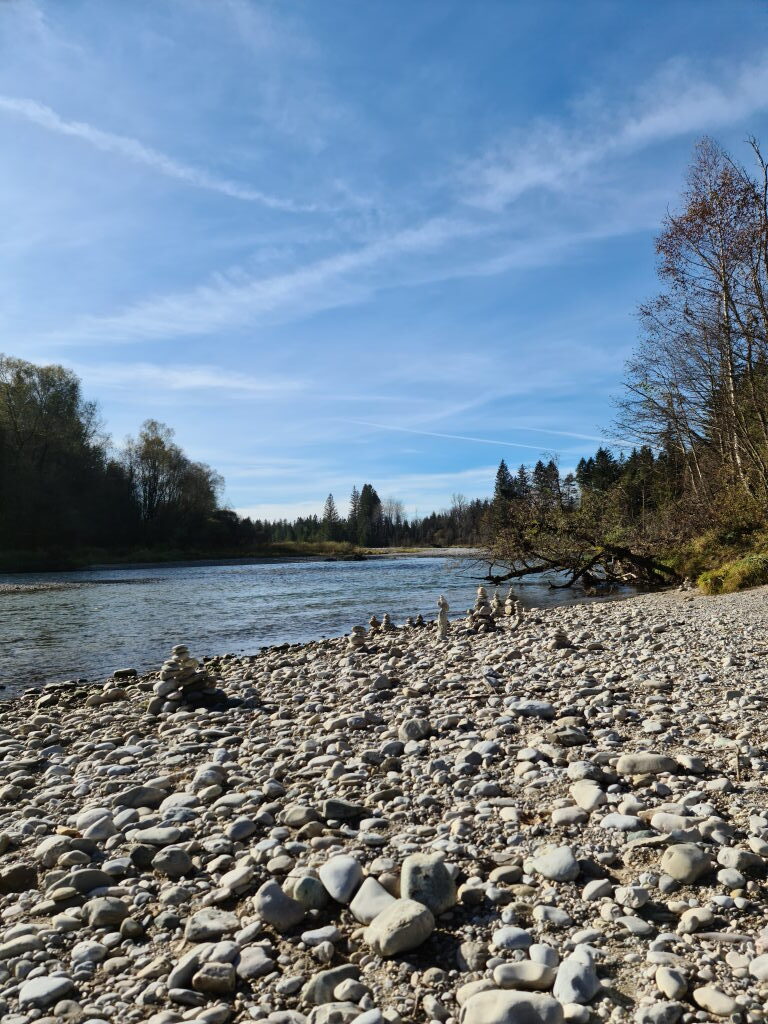 Steinmandln am Isarkiesufer bei Schäftlarn, gestapelte Kiesel, Fluss und Wald im Hintergrund