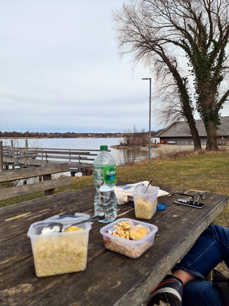 Picknick-Tisch am Chiemsee-Ufer mit Lunchboxen, Wasserflasche und Blick aufs Wasser, März 2021