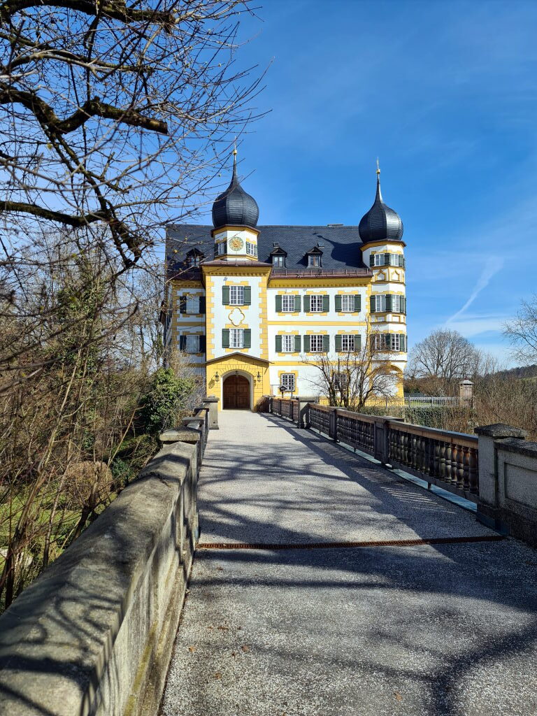 Schloss Wildenwart mit gelber Barockfassade und schwarzen Zwiebelturmhauben, Brücke zum Eingangstor, blauer Himmel, März 2021