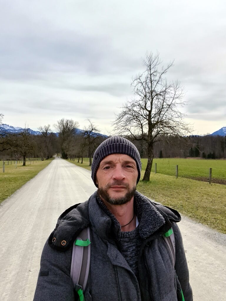 Sebastian als Wanderer in der langen Allee des Schlossparks Herrenchiemsee, Alpen im Hintergrund, März 2021