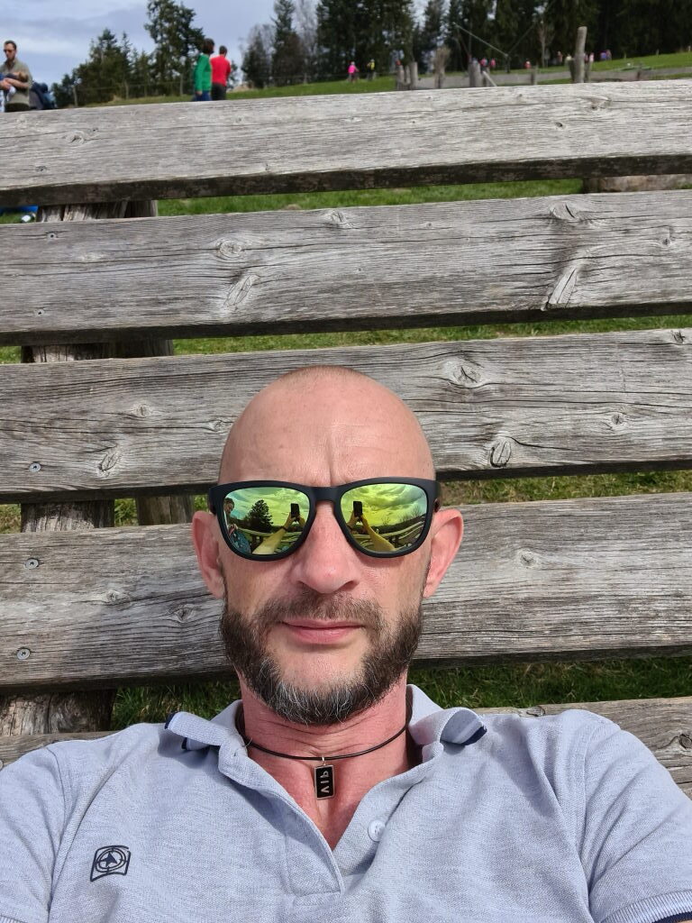 Sebastian auf einer Holzbank am Taubenberg mit Sonnenbrille und Blick in die Kamera