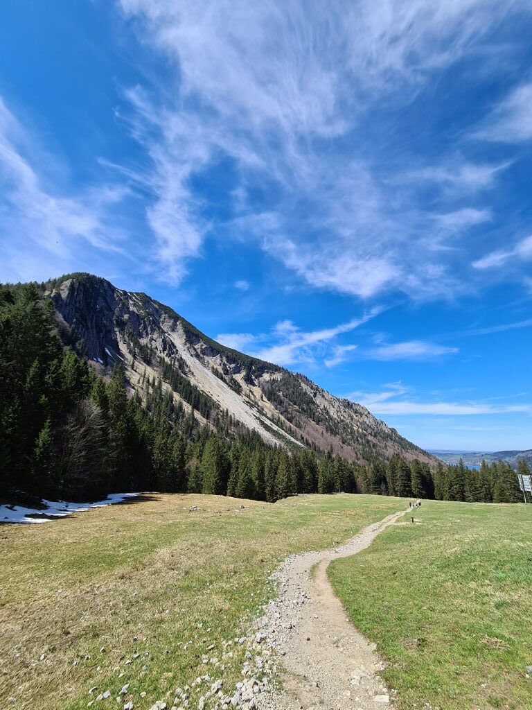 Schmaler Schotterweg über grüne Bergwiese mit mächtiger Felswand im Hintergrund und dramatischem Wolkenhimmel, Spitzingsee Wanderweg Mai 2021