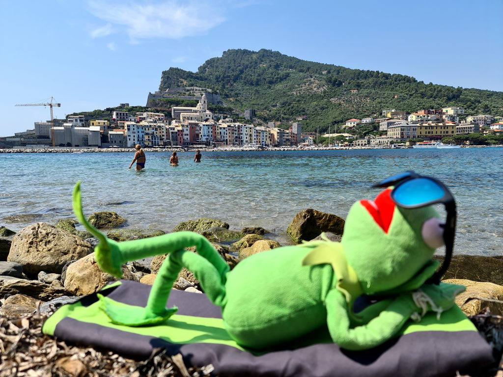 Kermit mit Sonnenbrille beim Sonnenbaden am Strand von Porto Venere mit Blick auf die Altstadt, Juli 2021