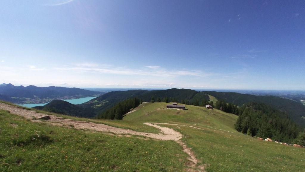 Weitläufige Almlandschaft unterhalb des Baumgartenschneid mit Almhütte, türkisem Tegernsee links und blauem Sommerhimmel