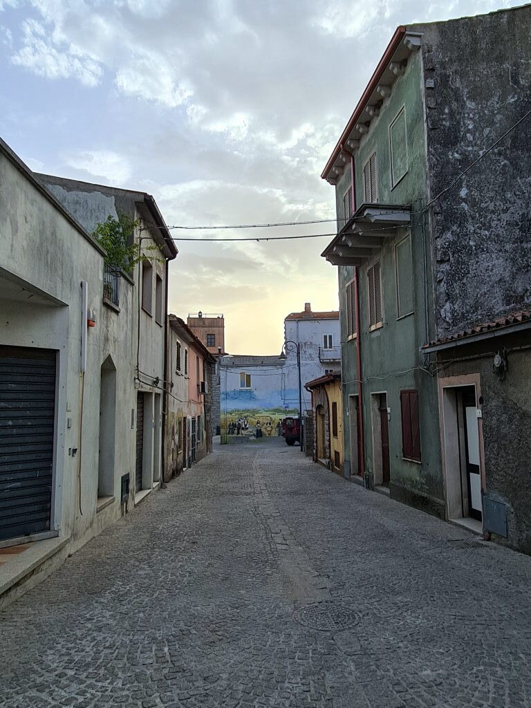 Kopfsteinpflastergasse in der Altstadt von Alà dei Sardi mit farbigen Häusern und einem Wandgemälde am Ende der Gasse