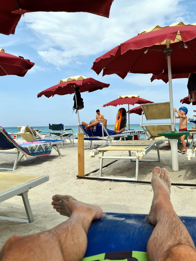 Strand in Budoni, Sardinien, mit burgunderroten Sonnenschirmen und Liegestühlen, Meer im Hintergrund