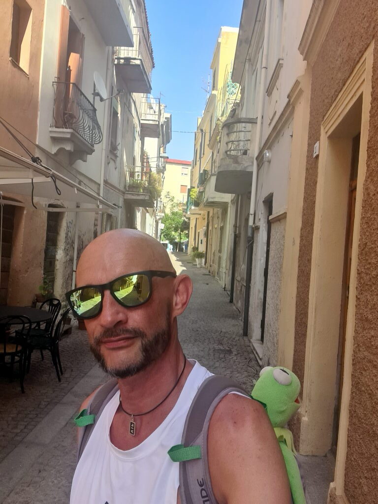 Sebastian Selfie in einer engen Altstadtgasse in Olbia, Kermit schaut über die Schulter aus dem Rucksack
