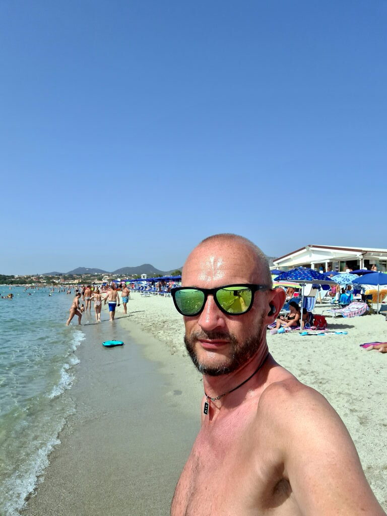 Sebastian Selfie an der Wasserlinie der Spiaggia Pittulongu, Sonnenbrillen, belebter Strand im Hintergrund