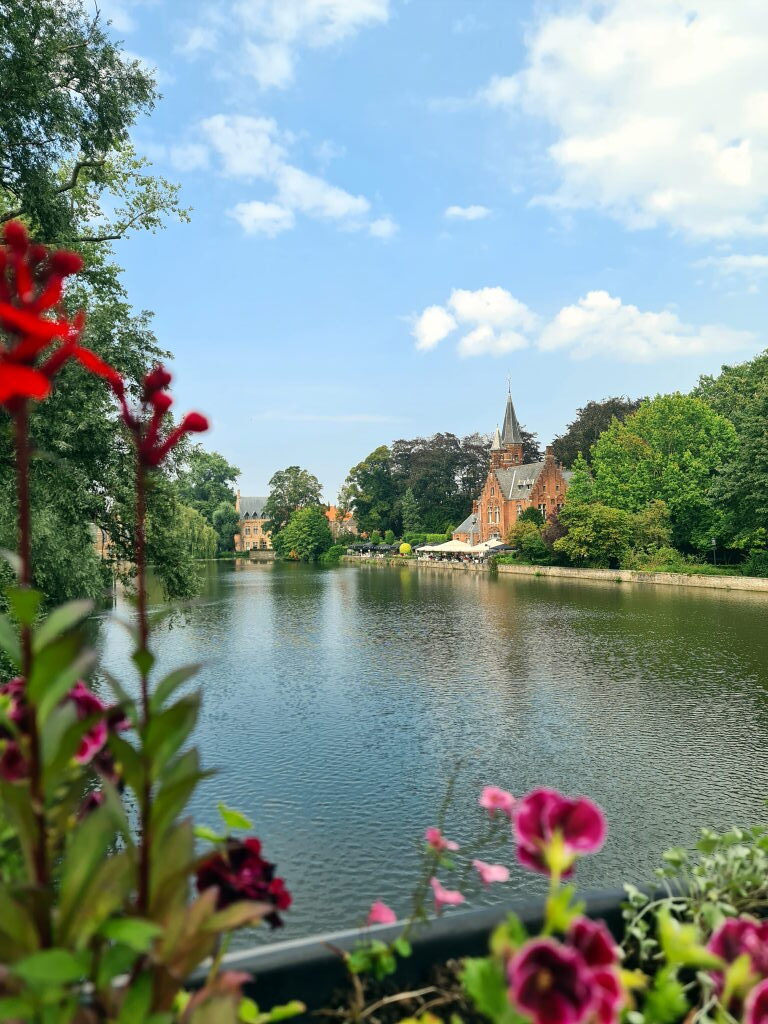 Blick über den Minnewater-See in Brügge mit dem roten Backsteinschloss im Hintergrund, umrahmt von Bäumen und bunten Blumen im Vordergrund
