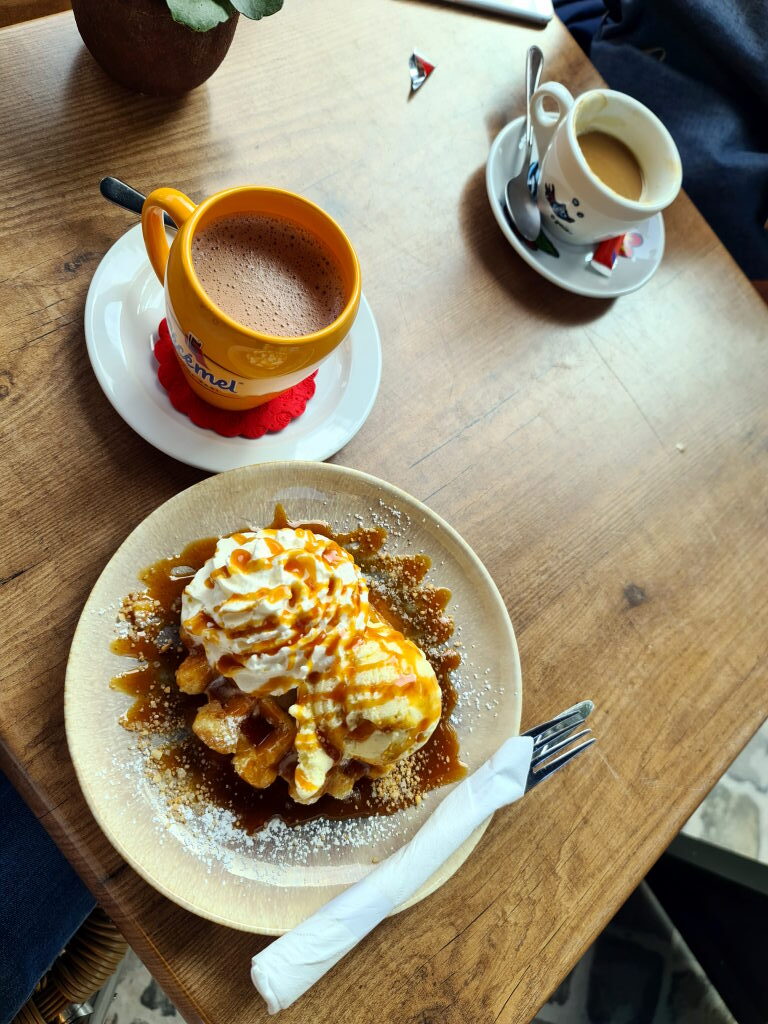 Belgische Waffel mit Schlagsahne und Karamellsauce auf einem Teller, daneben eine heiße Schokolade im gelben Kemelbecher und ein Espresso