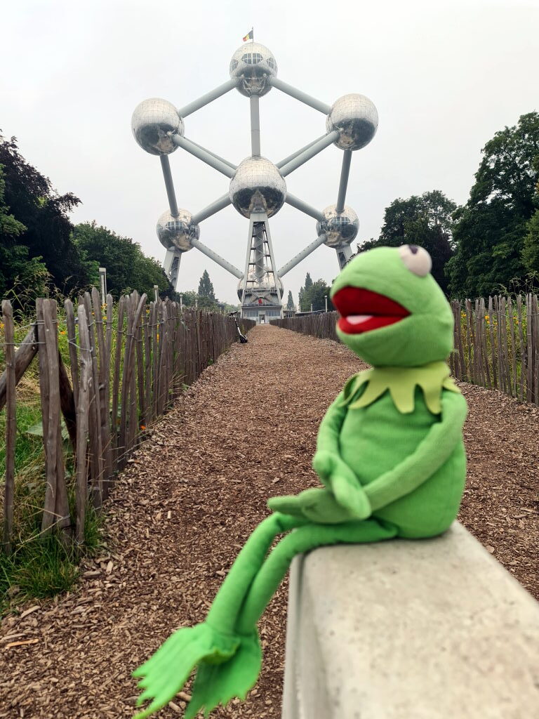 Kermit der Frosch sitzt auf einer Mauer vor dem Atomium in Brüssel, im Hintergrund die silbernen Kugeln des Bauwerks