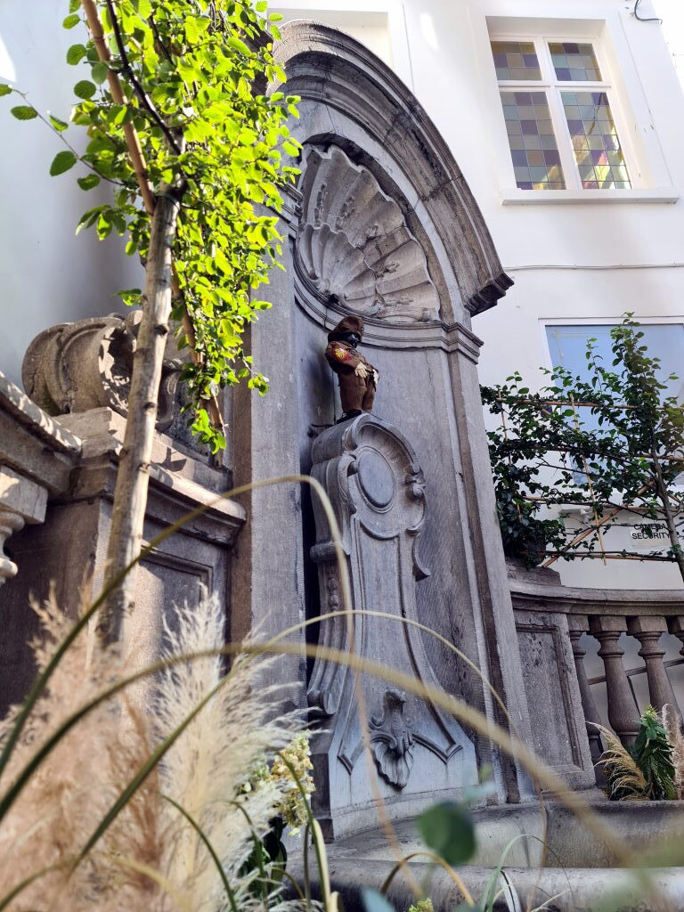 Der Manneken Pis Brunnen in Brüssel in einer barocken Steinnische, umgeben von Grünpflanzen, die Figur trägt ein buntes Kostüm