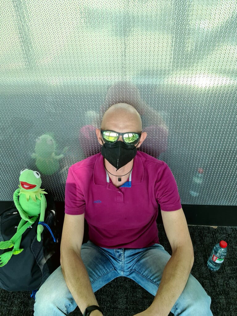 Sebastian mit schwarzer FFP2-Maske und Sonnenbrille sitzt am Brussels Airport, Kermit sitzt neben ihm am Rucksack