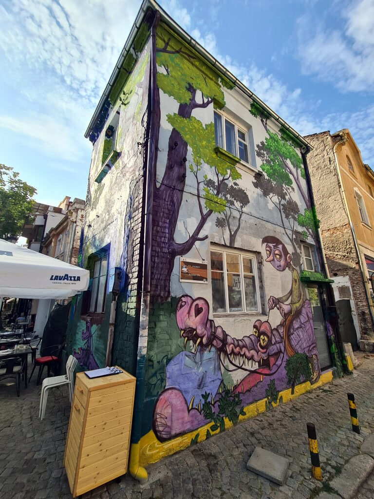 Mehrstöckiges Haus in der Plovdiver Altstadt komplett mit Street-Art-Mural bemalt, Junge auf Fantasietier, Bäume, Lavazza-Café-Schirm im Vordergrund