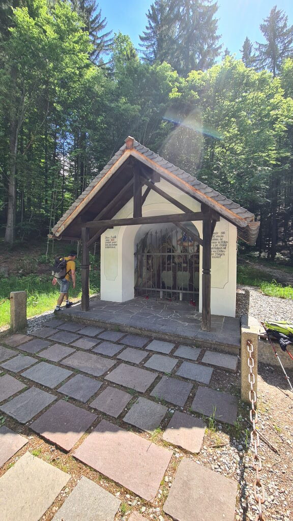 Kleine weiße Wegkapelle mit Holzdach und vergittertem Eingang im Wald bei der Geisterklamm