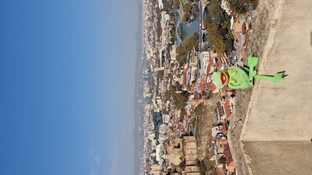 Kermit sitzt auf Mauer der Narikala-Festung mit Tiflis-Panorama und Friedensbrücke